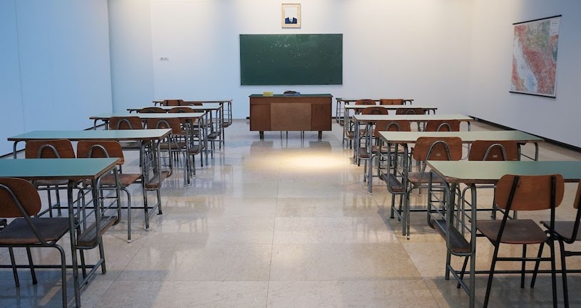 An empty classroom with a blackboard, teacher’s desk, and rows of desks and chairs.
