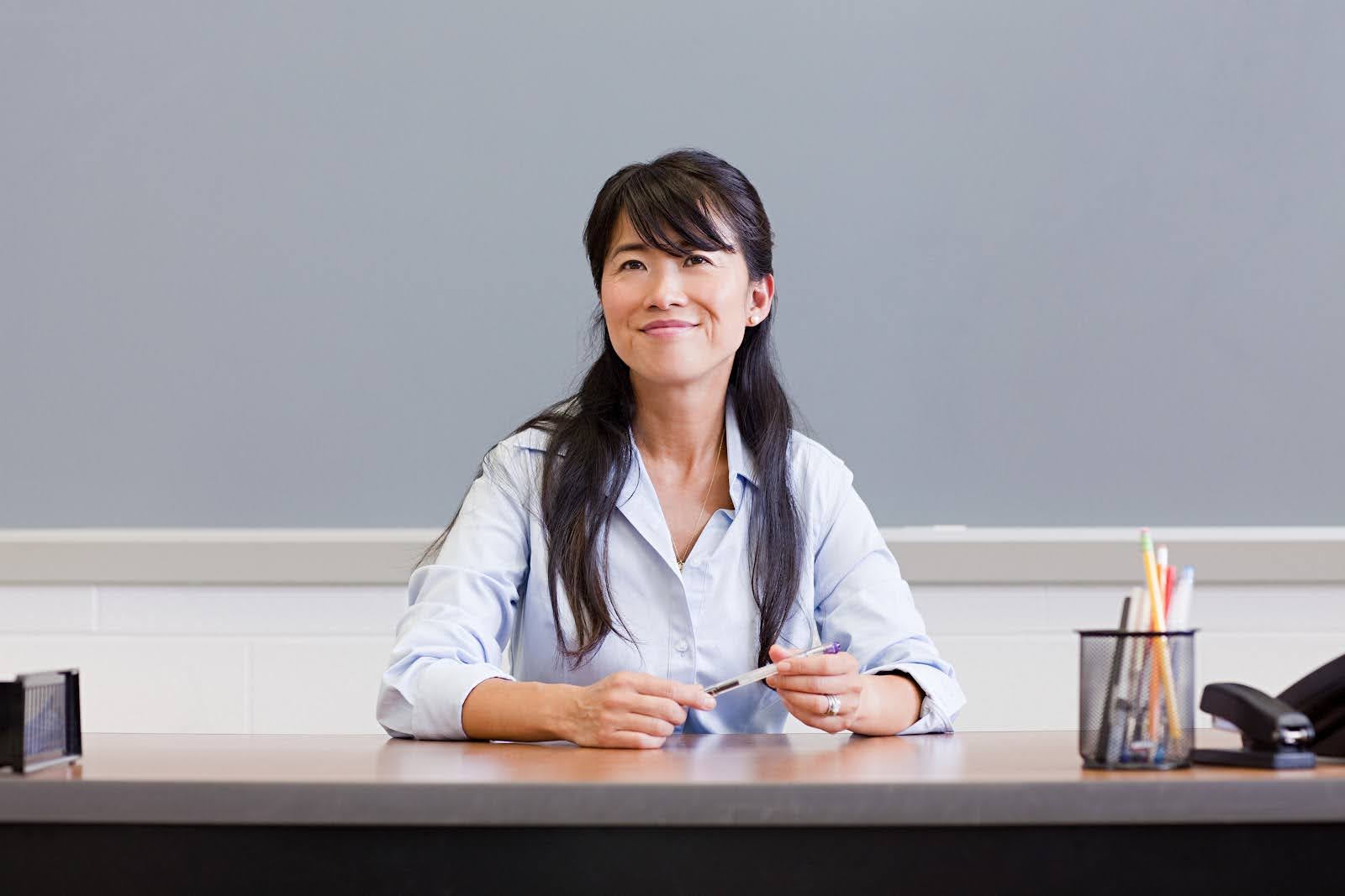 A smiling teacher holding a pen sits at her desk; in the foreground we see a stapler, a document organizer, and a container of pens and pencils.