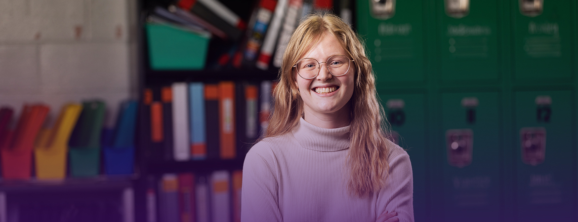 A person with glasses and long hair smiles, standing in front of green lockers and shelves with files and binders.