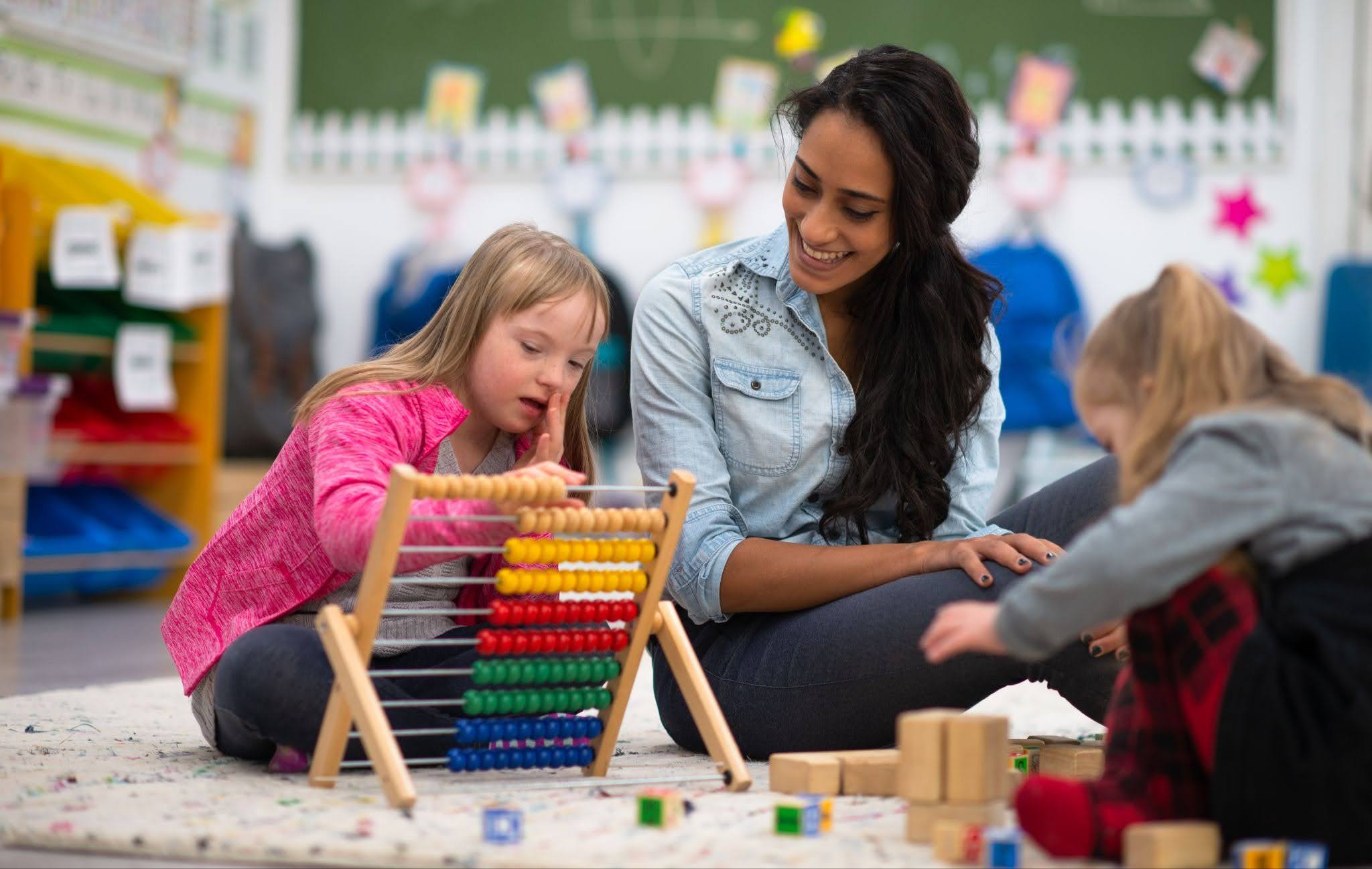A young girl in a classroom setting uses an abacus while a smiling teacher observes.