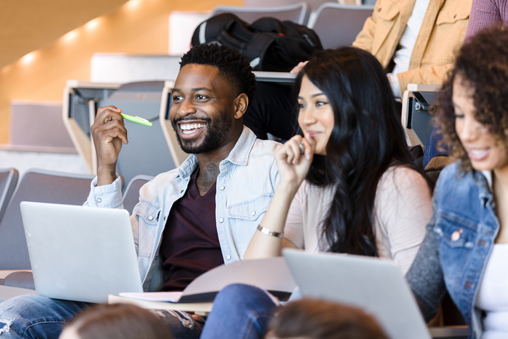 Smiling students seated in a lecture hall, engaging with laptops open and pens in hand.