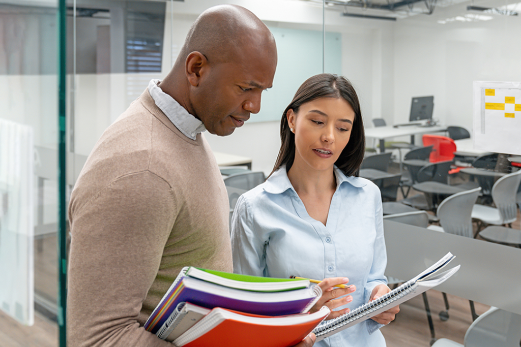 Two people standing in a classroom, reviewing notes and holding notebooks while having a discussion.