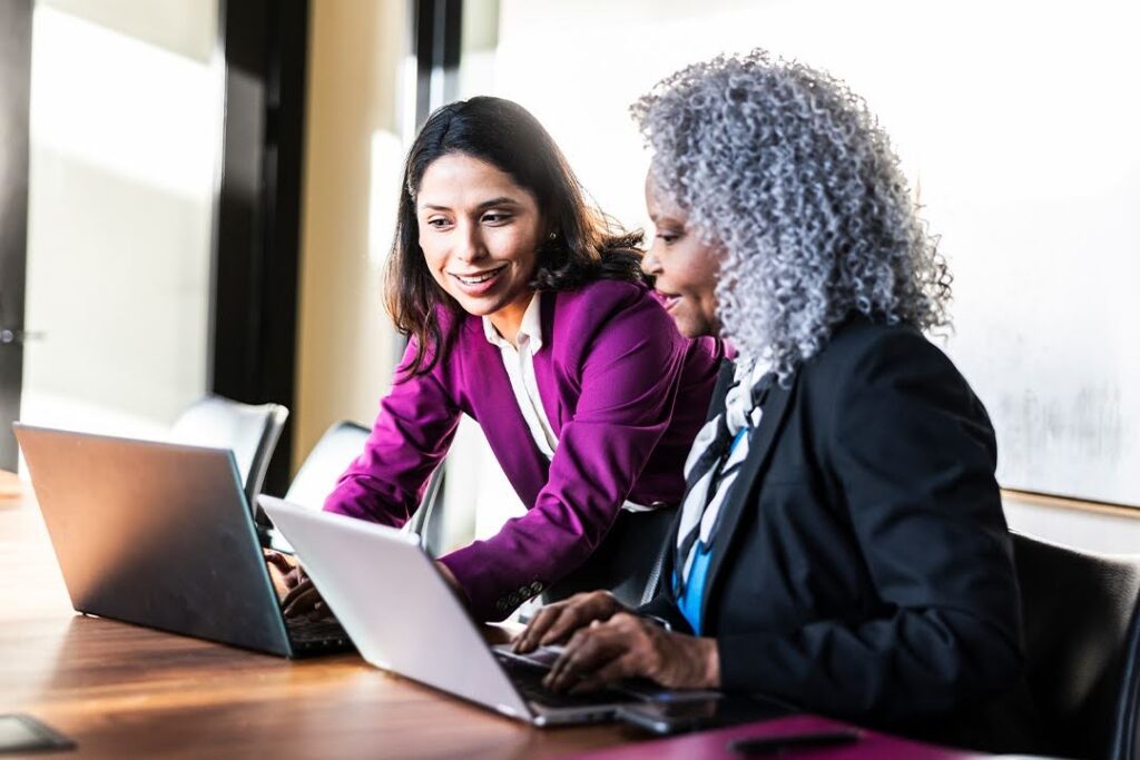 Two smiling women in business attire converse and work together at laptops; the setting is a modern office space.