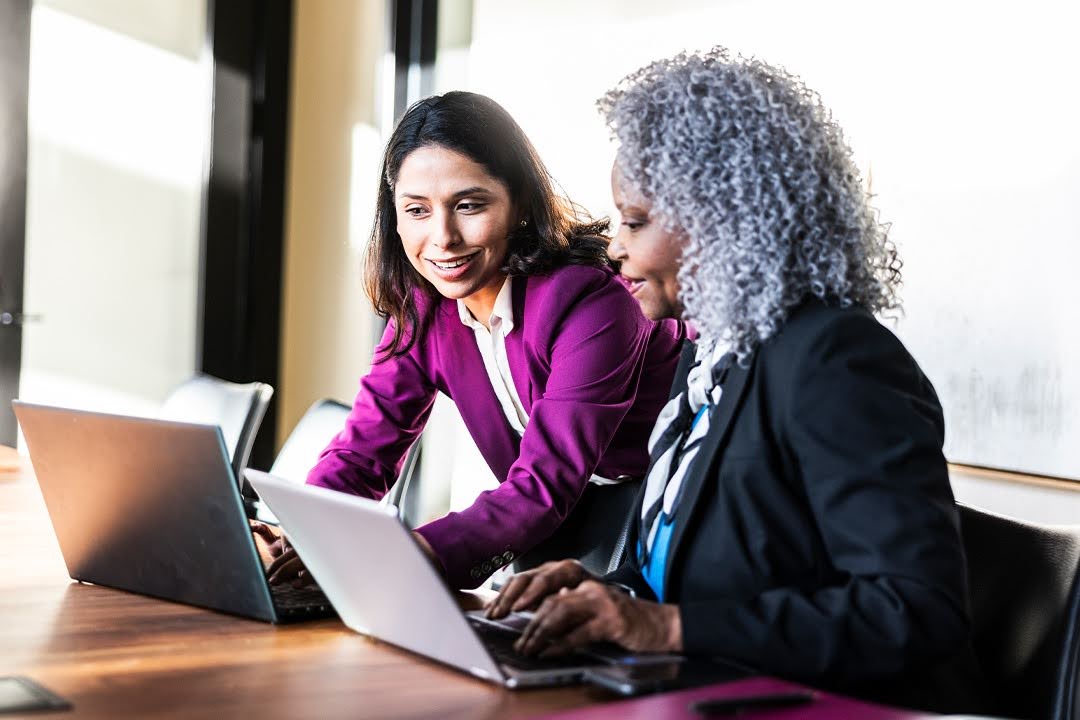 Two smiling women in business attire converse and work together at laptops; the setting is a modern office space.