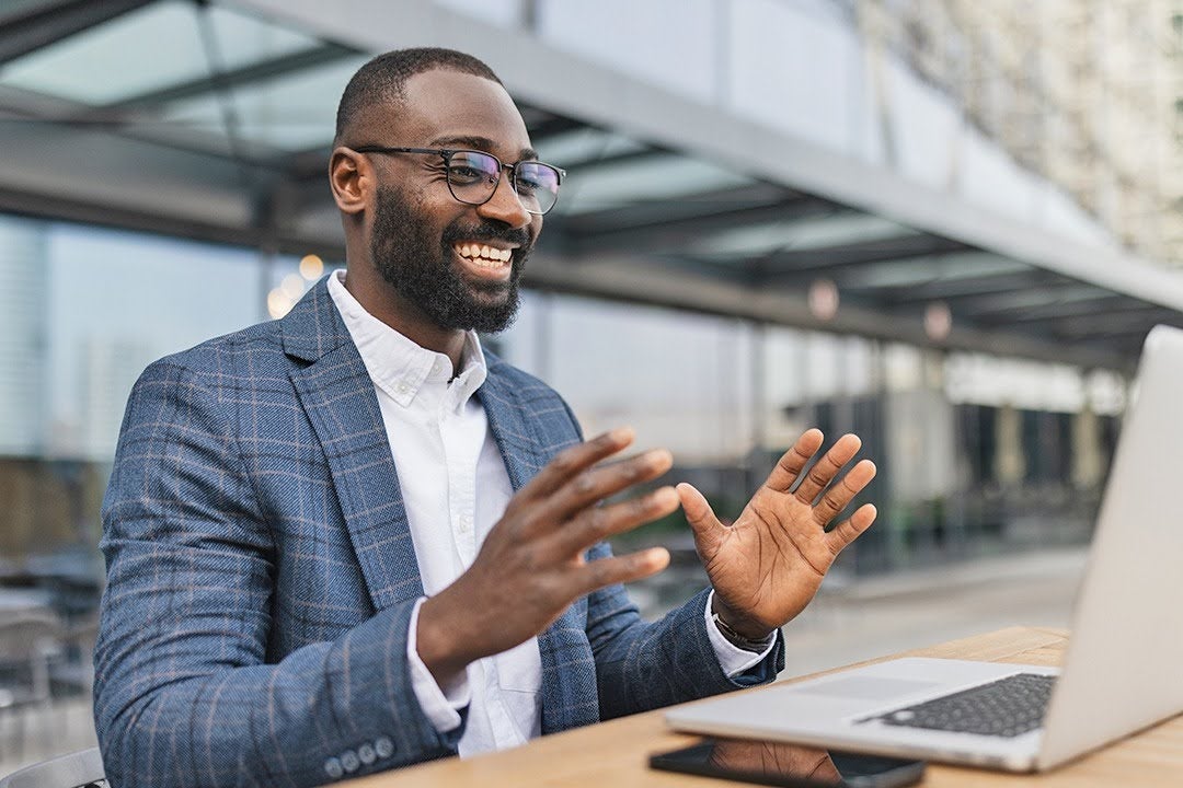 A smiling man in a blazer and glasses gestures while video-chatting on a laptop at an outdoor table; a modern glass building can be seen in the background.