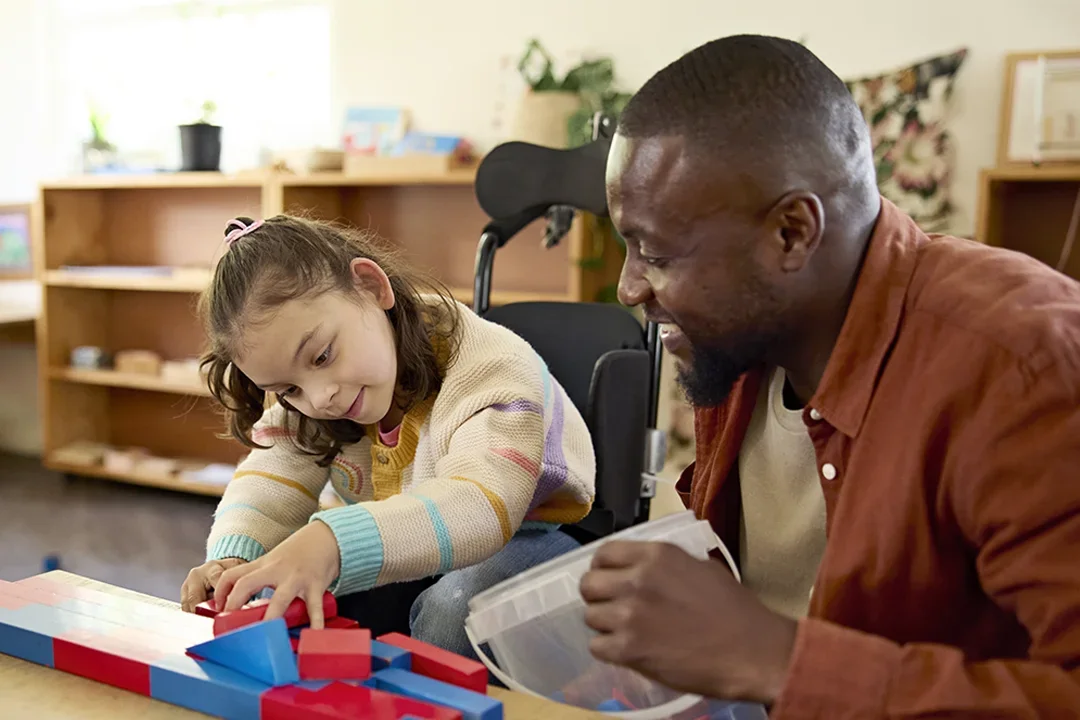 A teacher smiles as he overlooks a child playing in a wheelchair.