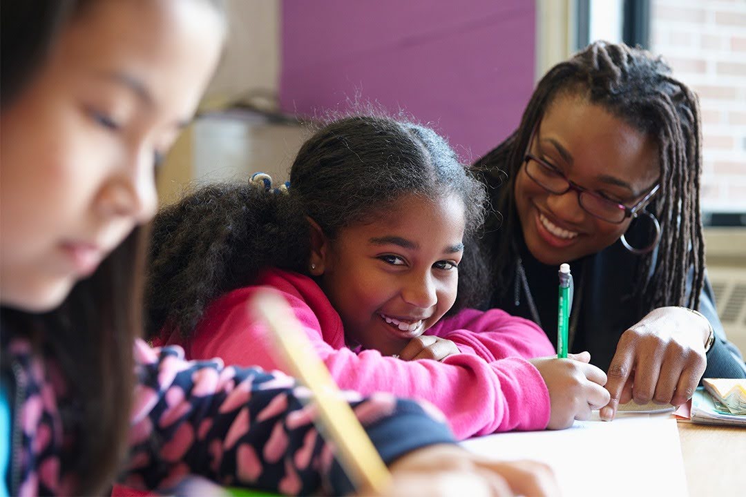 A smiling teacher guides a young girl writing at her desk in a classroom.